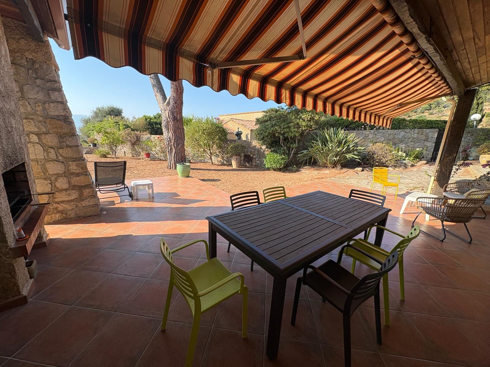 Terrasse de la villa de Sagone sous le store, grande table pour huit, barbecue en pierre et jardin